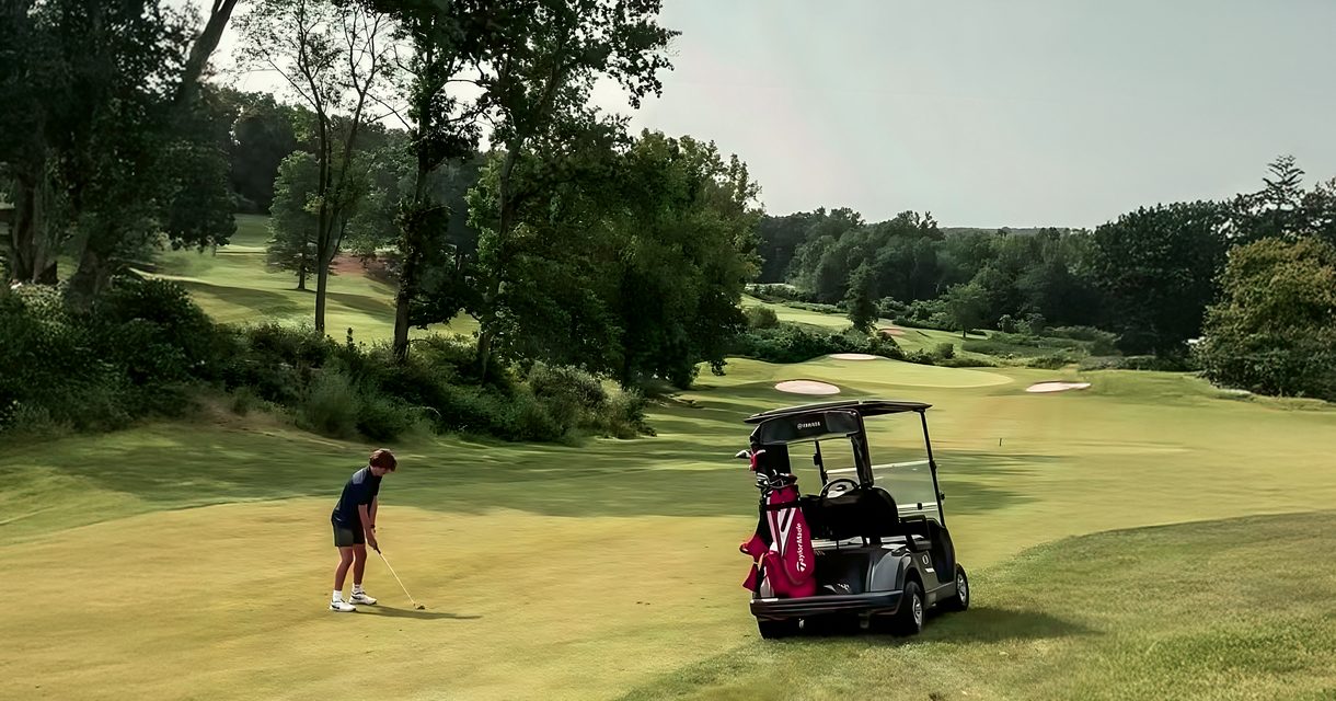 Golfer chipping toward the green beside a golf cart on a tree-lined fairway at The VUE