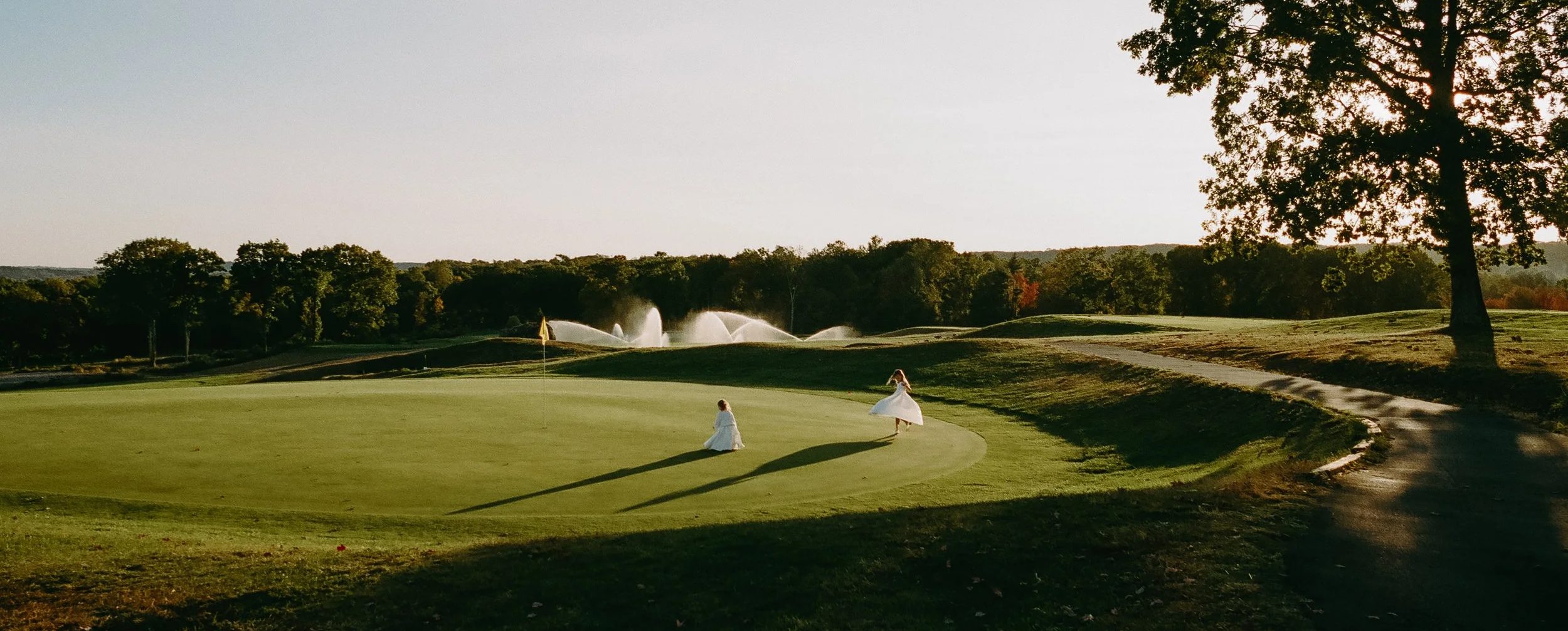 Two women in white wedding gowns twirling on the green of The VUE golf course at golden hour
